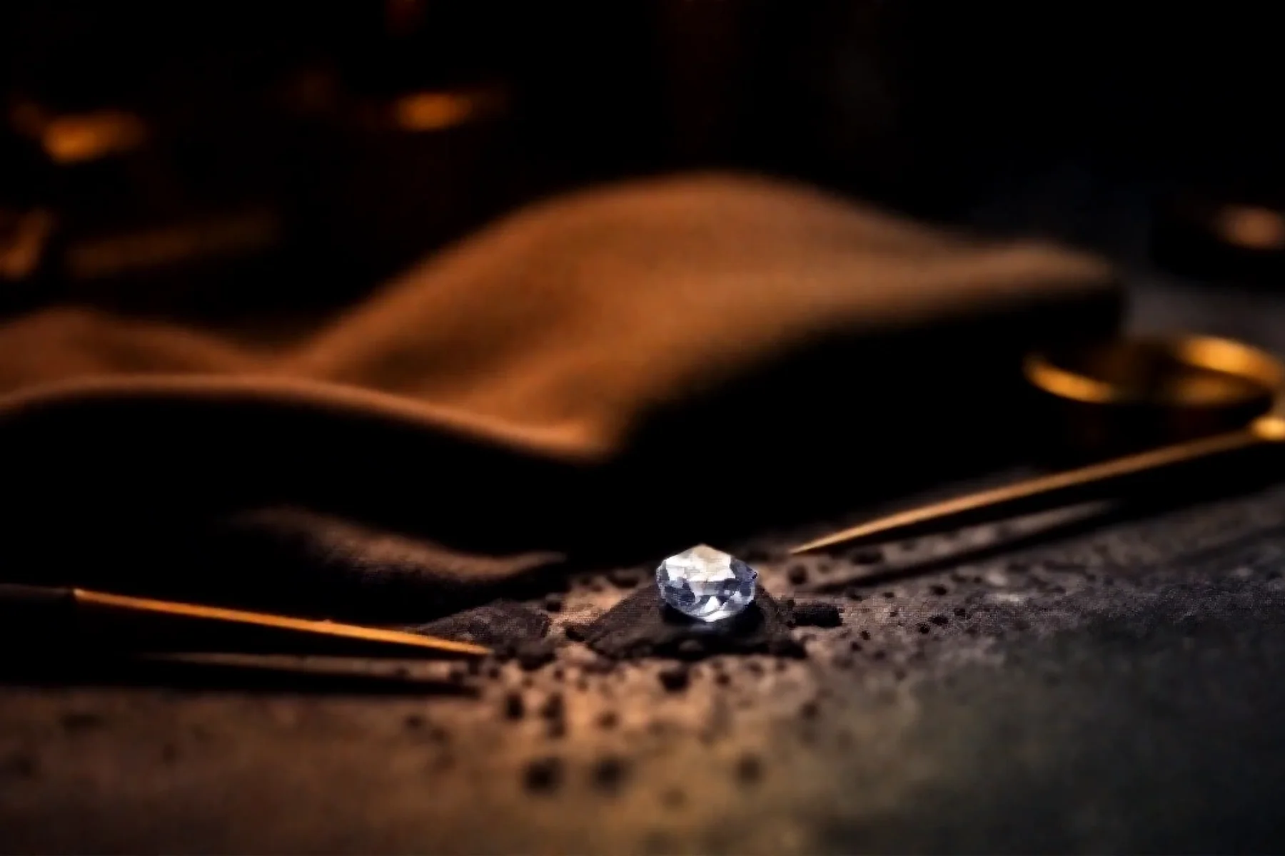 A loose diamond, tweezers, and a jeweler's loupe on a dark workbench